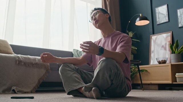 Ground level shot of young Asian man in casualwear meditating in lotus pose on floor and turning on relaxing music on mobile phone in living room