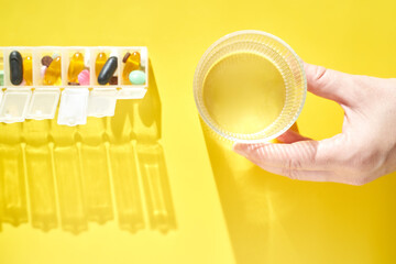 Hand holding glass of water next to weekly pill organizer on bright yellow background in a health and wellness setting.
