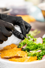 Close-up of gloved hands plating breaded chicken cutlets with fresh green salad and radishes on a white plate in a kitchen setting, emphasizing hygiene and food presentation.