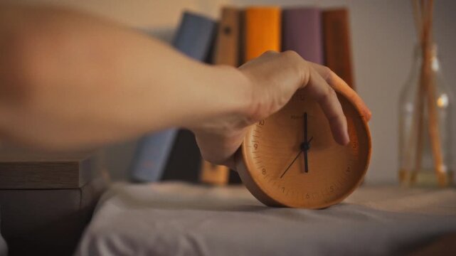 Close-up of man's hand reaching for an alarm clock turning off the alarm in the early morning. An analog clock stands on small table against the background of books. The man turns off the alarm clock