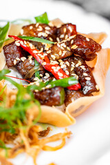 Close-up of glazed beef with sesame seeds, chili, and green onion in a crispy edible bowl, served on a white plate. Gourmet Asian dish with vibrant colors and fine presentation