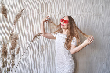 Caucasian young female in white lace dress with red sunglasses posing by textured wall with dried...