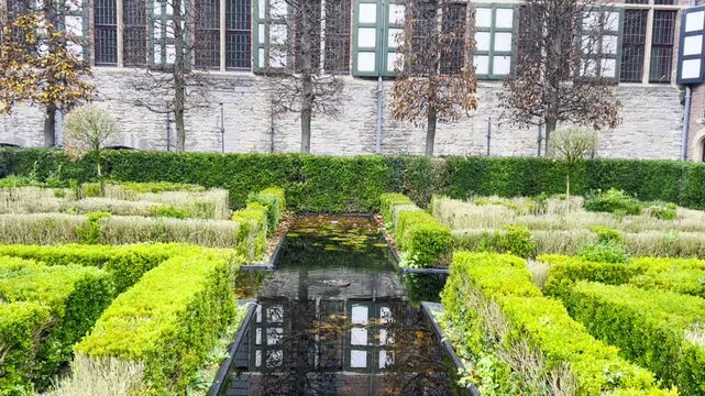 Stunning maze garden features reflecting pool at its center, surrounded by lush greenery and historic brick buildings, creating a serene oasis perfect for relaxation and contemplation. Gent, Belgium