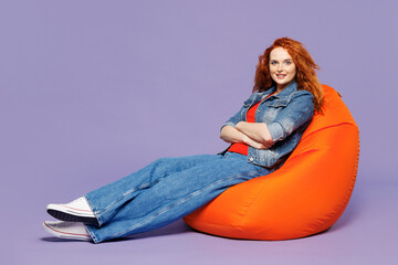 Full body side view young ginger woman she wears red t-shirt denim shirt sit in bag chair hold...