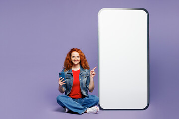 Full body young ginger woman she wears red t-shirt denim shirt sit big huge blank screen mobile cell phone smartphone point finger aside on area isolated on plain purple background. Lifestyle concept.