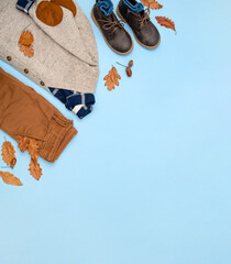 Children&rsquo;s autumn outfit flat lay with sweater, plaid shirt, brown shorts and leather boots, decorated with oak leaves and acorns on blue background. Cozy fall wardrobe concept. Top view, copy space.