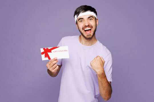 Young winner strong fitness trainer sporty man sportsman wear headband t-shirt train in gym hold gift coupon voucher card for store isolated on plain purple background. Workout sport fit abs concept.