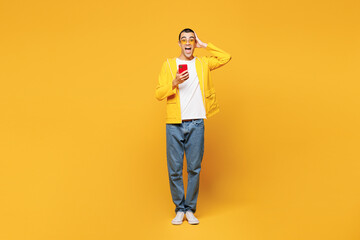 Full body young shocked surprised middle eastern man he wear raincoat casual clothes hold in hand use mobile cell phone isolated on plain yellow orange background studio portrait. Lifestyle concept.