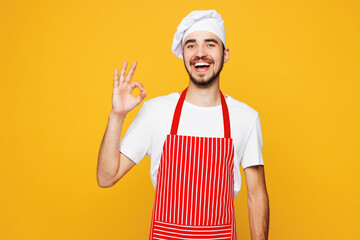 Young smiling happy fun housewife housekeeper chef cook baker man wearing red apron toque hat showing okay ok gesture isolated on plain yellow orange background studio portrait. Cooking food concept.