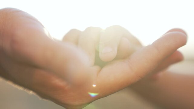 Hands holding child hand gently under sunlight with sun lensflare and soft focus showing palm touch and intimate connection between parent and child closeup warmth tenderness care glow hope bonding