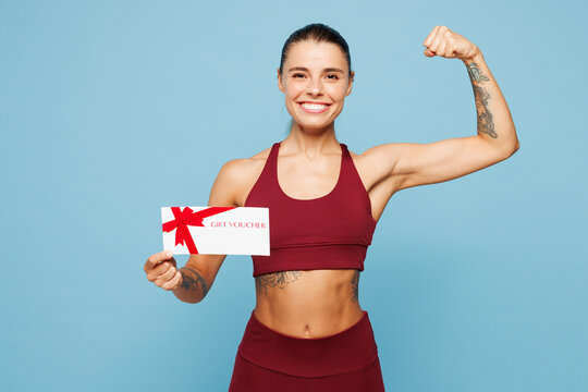 Young fitness trainer instructor sporty woman in red sportswear train in home gym hold gift coupon voucher card for store show muscles isolated on plain blue background Workout sport fit abs concept