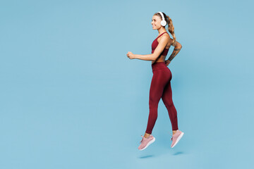 Fototapeta premium Full body young fitness trainer instructor sporty woman sportsman wear red sportswear train in home gym listen music in headphones jump isolated on plain blue background Workout sport fit abs concept