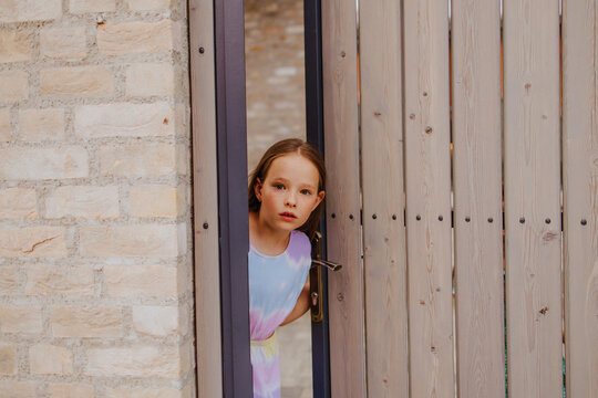 Child peeking out through wooden gate at home with scared expression