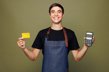 Young man barista bartender barman he wear apron t-shirt work in coffee shop hold bank payment terminal process acquire credit card isolated on plain green background. Small business startup concept.