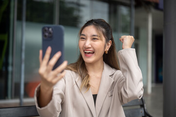 Asian woman holding phone raising a fist looking excited while sitting on bench in front of a store.