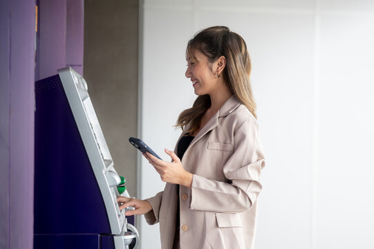 Smiling asian woman standing holding phone in front of an atm machine waiting for a withdraw money.