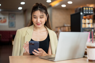 Smiling asian woman holding and looking at phone aside laptop while sitting at wooden table in cafe.