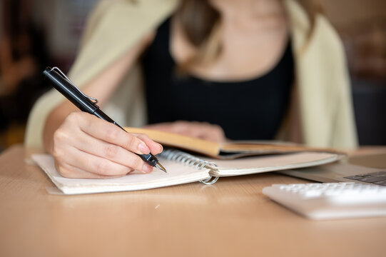 Close up of woman's hand holding pen writing in notebook aside calculator and laptop on wooden table