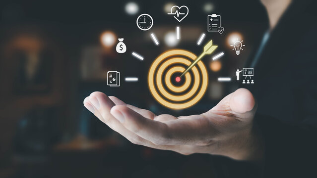 A person's hand holds a glowing target with an arrow,surrounded by icons for goals like finance, health,and time. This illustrates the concept of setting and achieving personal or business objectives.