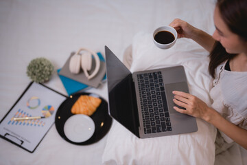 Businesswoman is working from home in bed, drinking coffee and eating a croissant, using laptop and analyzing statistics, enjoying breakfast