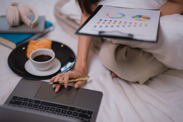 Businesswoman is comfortably working from bed, analyzing charts and using laptop while enjoying a...