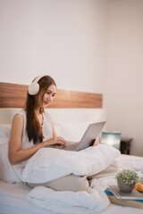 Freelancer is working from home, sitting comfortably in bed with pillows and using a laptop and headphones, enjoying the flexibility and comfort of remote work