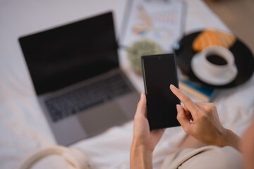 Freelancer woman is using smartphone with laptop and breakfast on bed in hotel room enjoying remote work and travel lifestyle combining business and leisure