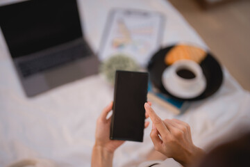 Woman lying in bed, using smartphone with blank screen, surrounded by laptop, breakfast, and documents on the sheets, enjoying a cozy morning while working from home or relaxing