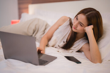 Young woman lying on a cozy bed, working on a laptop with a smartphone nearby, engaging in online studies and enjoying the comfort of her home environment during the evening