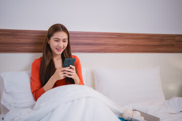 Happy asian woman is using smartphone while relaxing in bed in the morning, she is using smartphone for social media, shopping online and chatting with friends