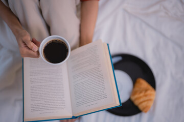 Woman sitting in bed, engrossed in a book while savoring a cup of coffee and a flaky croissant on a...