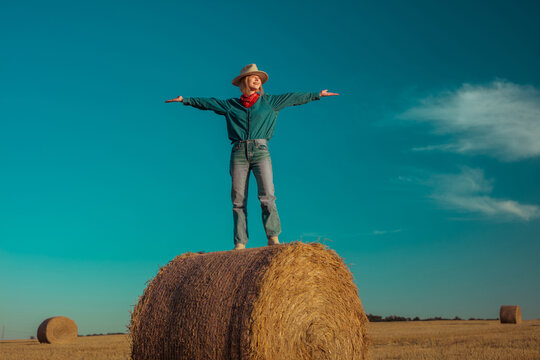 Stylish cowgirl standing on hay bale in wheat field at sunset