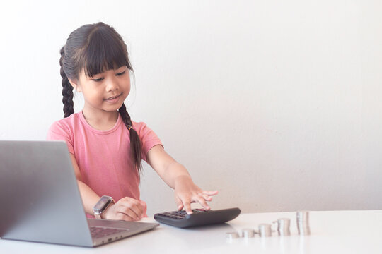 A cute Asian girl is using a calculator to calculate some statistics. There is a pile of coins and a laptop on the table.