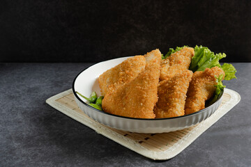 Risol Mayo segitiga or Triangle Risol mayonnaise served in a white bowl with fresh lettuce and green chili on dark background. Showcasing a savory Indonesian snack with breadcrumb coating. 