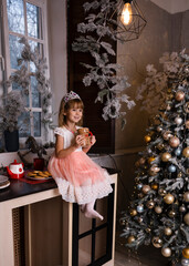 A girl in a pink dress sits alone on a table and drinks cocoa near a New Year tree.