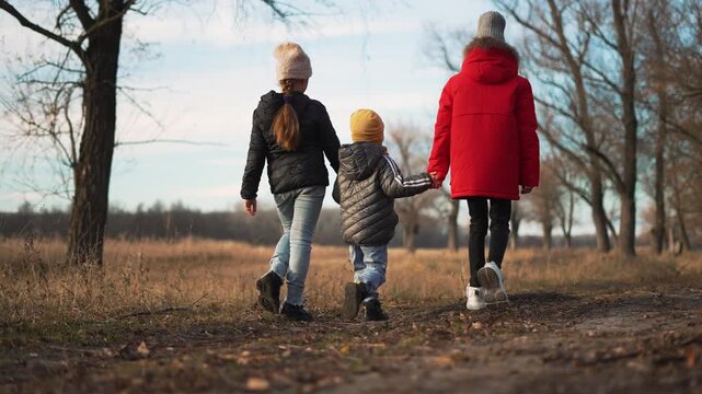 Three children walk together holding hands along muddy path across frost field and bare tree row in park wearing winter jacket and hat displaying family bond and sibling care with child playful energy