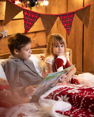 A boy and girl enjoy reading a book on a bed surrounded by a cozy Christmas atmosphere with lights and seasonal decor.