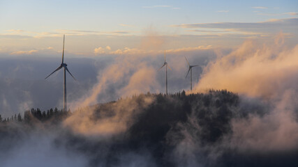 Aerial view of wind turbines piercing through a sea of ethereal clouds, their stark silhouettes contrasting against the soft, pastel sky, Gaberl, Styria, Austria.