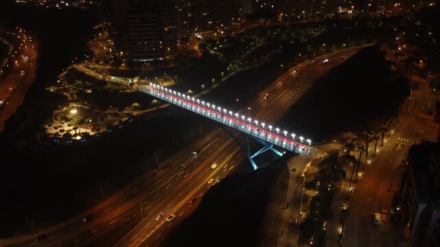 Night time aerial footage of a bridge connecting two districts of Miraflores and Barranco, located in Lima, Peru. The bridge hangs over a street named "Bajada Armendariz". Drone hovers and tilts up.