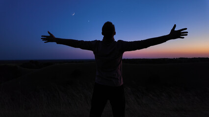 Silhouette of a man and countryside under the Moon, stars and planets in blue hour time.