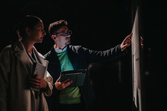 Two people engaged in discussion, attentively observing a display screen in a dimly lit context, evoking themes of analysis, teamwork, and appreciation for knowledge or art in an immersive setting.