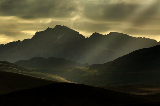 View of sun rays piercing through the dramatic, moody clouds over the rugged, dark mountain ranges, creating a serene yet powerful scene, Deosai National Park, Gilgit Baltistan, Pakistan.