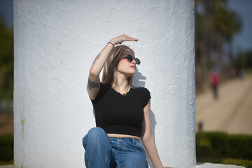 Young woman, beautiful, blonde, wearing a black t-shirt, jeans, and sunglasses, sitting on white...