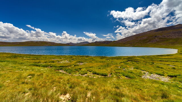 View of the calm, azure lake mirroring the sky's fluffy clouds, embraced by vibrant green meadows and rugged mountains, Deosai National Park, Gilgit Baltistan, Pakistan.