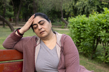 unhealthy indian woman sitting alone on bench look down feel dull headache touching head brain at...