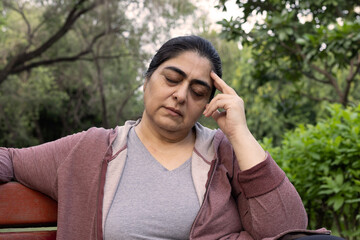 unhealthy indian woman sitting alone on bench look down feel dull headache touching head brain at...