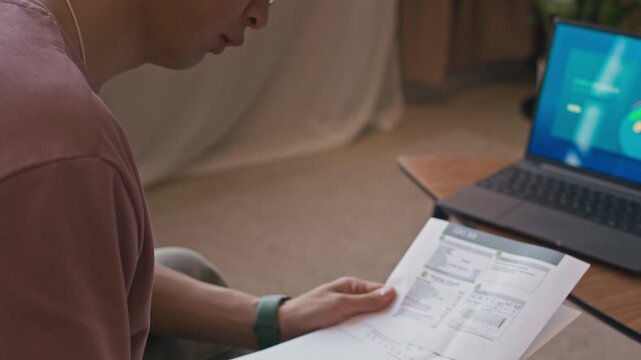 Tilt up shot of young man calculating expenses while checking invoices sitting in front of laptop in home office