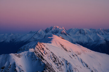 Naklejka premium Aerial view of snow-covered mountain peaks bathed in soft pink and mauve light, highlighting jagged ridgelines and textured rock under an expansive early morning or late evening sky.