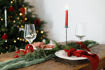 Merry Christmas! Stylish table setting for christmas family dinner. Modern plate and cutlery with napkin, glassware, green and red ribbons, fir branches and candle on rustic table at xmas tree