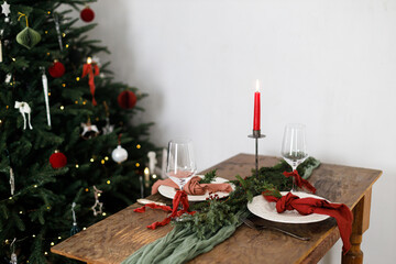 Stylish christmas table setting for family dinner. Elegant plate with napkins, cutlery, glassware, ribbons, green fir branches, red berries and candle at xmas tree lights. Holiday time